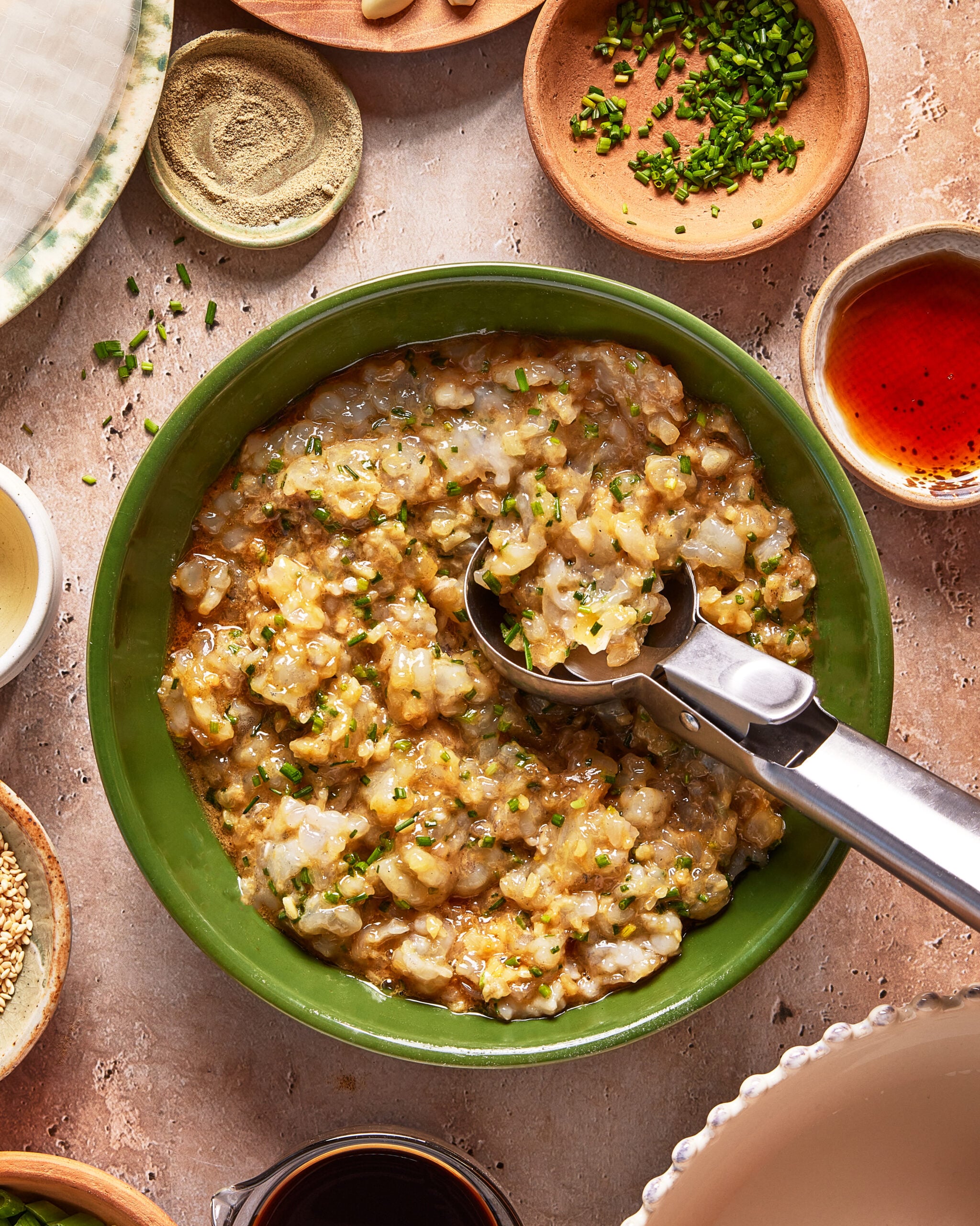 A green bowl filled with a chunky mixture, garnished with chopped chives, with a metal scoop resting inside. Surrounding the bowl are small dishes containing sauces, spices, and herbs.