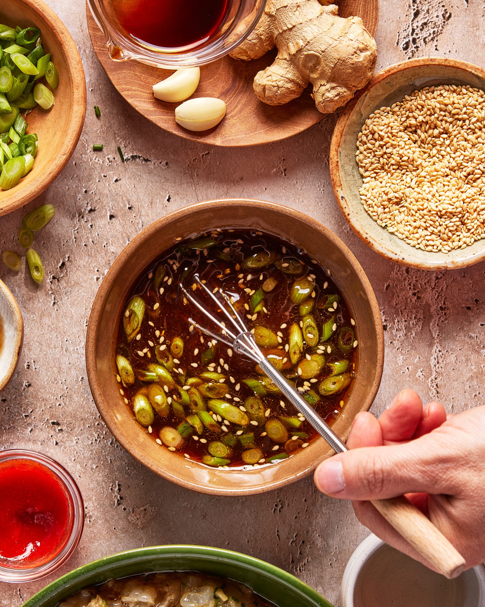 A hand whisking a brown sauce with chopped green onions in a bowl, surrounded by sesame seeds, ginger, garlic, green onions, and other sauce ingredients on a textured surface.