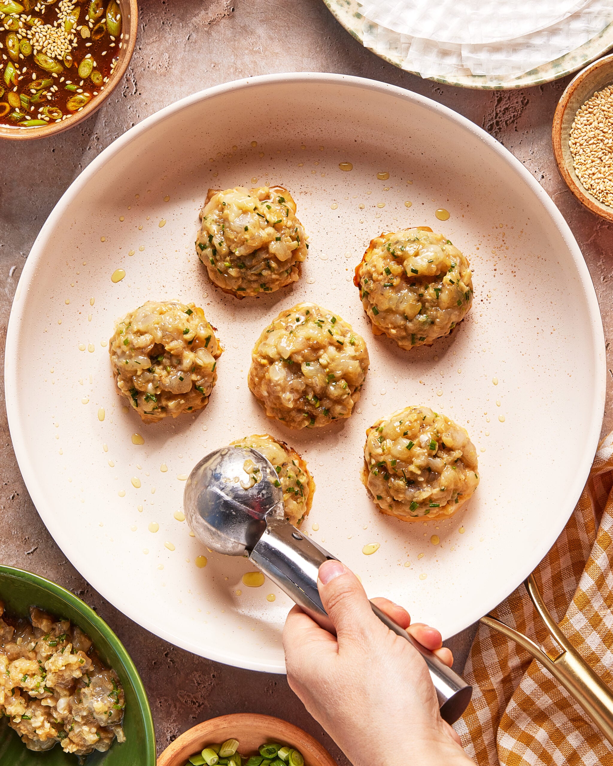 A hand uses an ice cream scoop to portion out five round scoops of a vegetable and meat mixture onto a white plate, with bowls of sauce, sesame seeds, and green onions nearby.