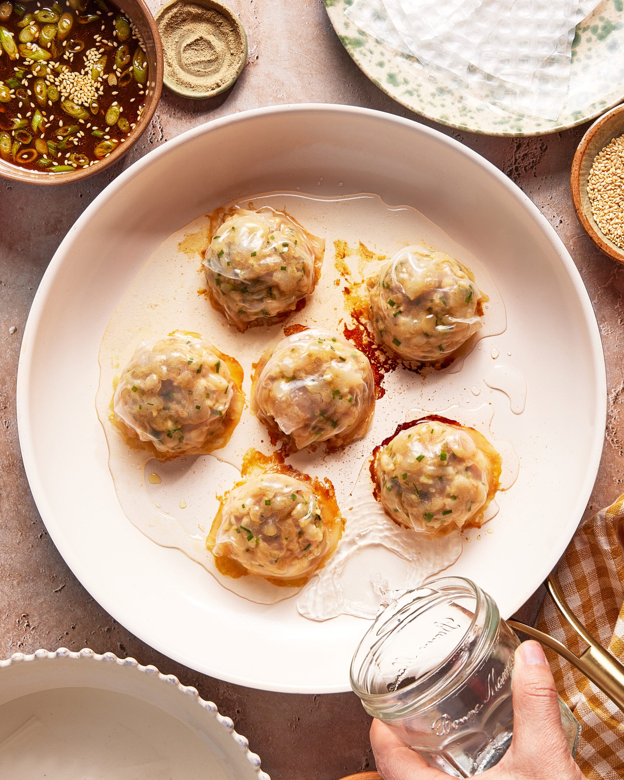 A hand holding a glass jar pours water onto a plate with six translucent dumplings filled with herbs and meat. Nearby are dishes of dipping sauce, sesame seeds, and rice paper wrappers.
