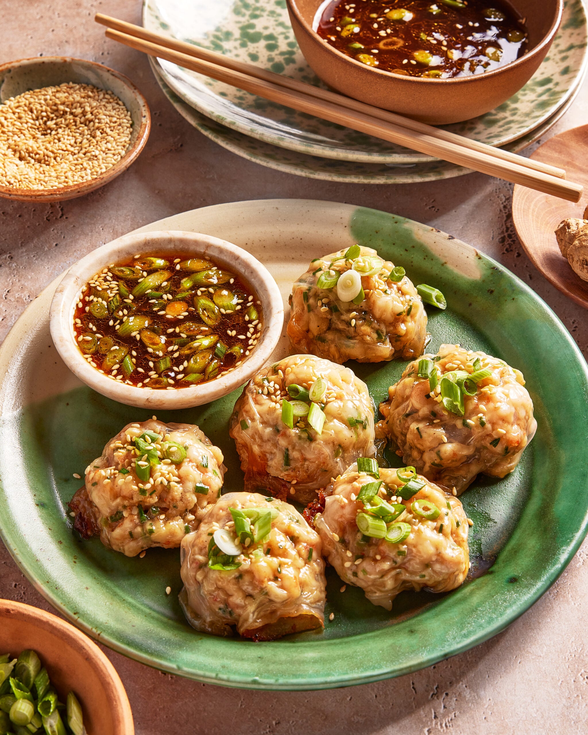 A green plate with six steamed dumplings garnished with chopped green onions and sesame seeds, served with a small bowl of dipping sauce, surrounded by dishes of sauce and sesame seeds. Chopsticks rest nearby.