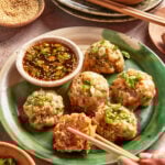 A green plate with six steamed dumplings topped with chopped green onions, served with a bowl of soy dipping sauce. A hand with chopsticks picks up one dumpling. Small bowls of sesame seeds and sauce are nearby.