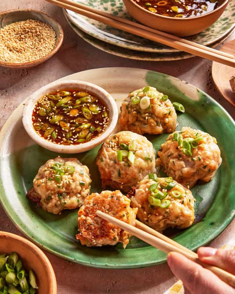 A green plate with six steamed dumplings topped with chopped green onions, served with a bowl of soy dipping sauce. A hand with chopsticks picks up one dumpling. Small bowls of sesame seeds and sauce are nearby.