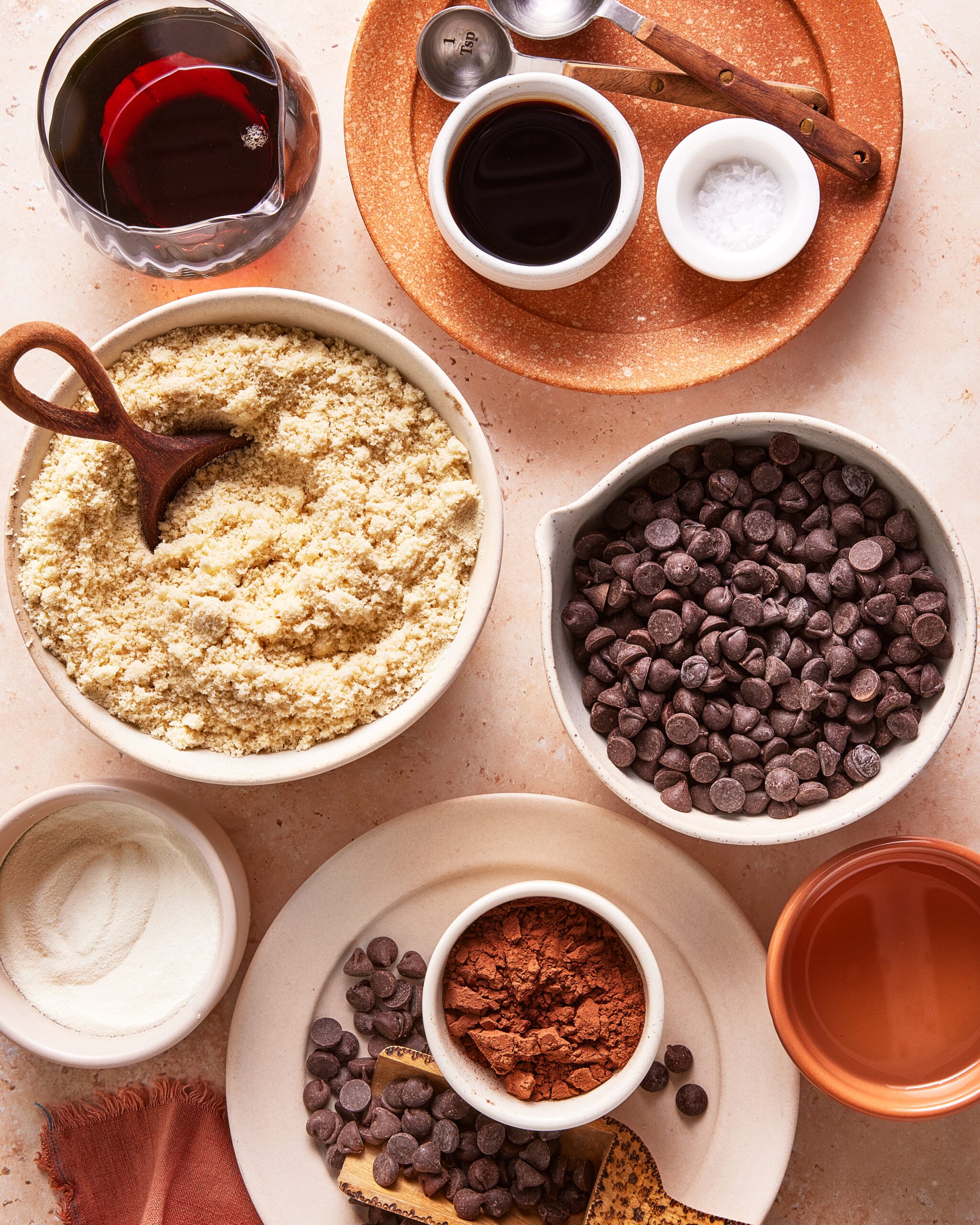An overhead view of small bowls filled with baking ingredients: almond flour, chocolate chips, cocoa powder, sugar, vanilla extract, salt, and a glass of red wine, arranged neatly on a light countertop.