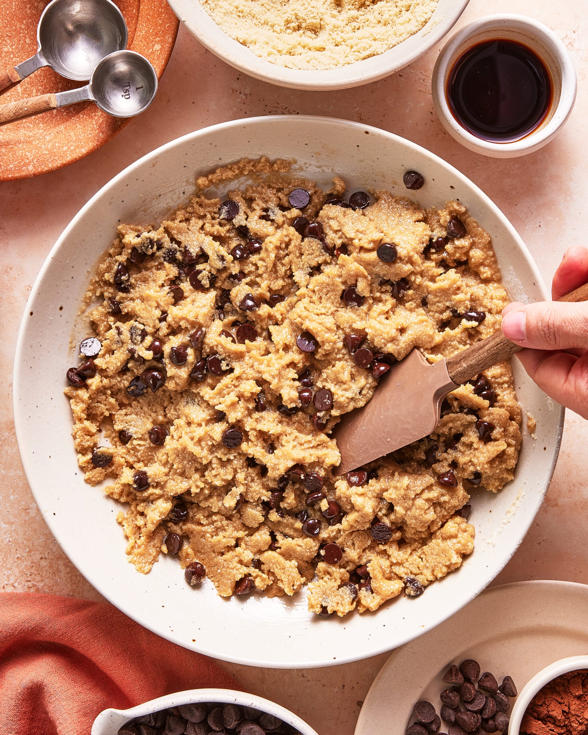 A hand stirs chocolate chip cookie dough in a white bowl with a spatula. Surrounding the bowl are small dishes containing chocolate chips, flour, vanilla extract, and measuring spoons.
