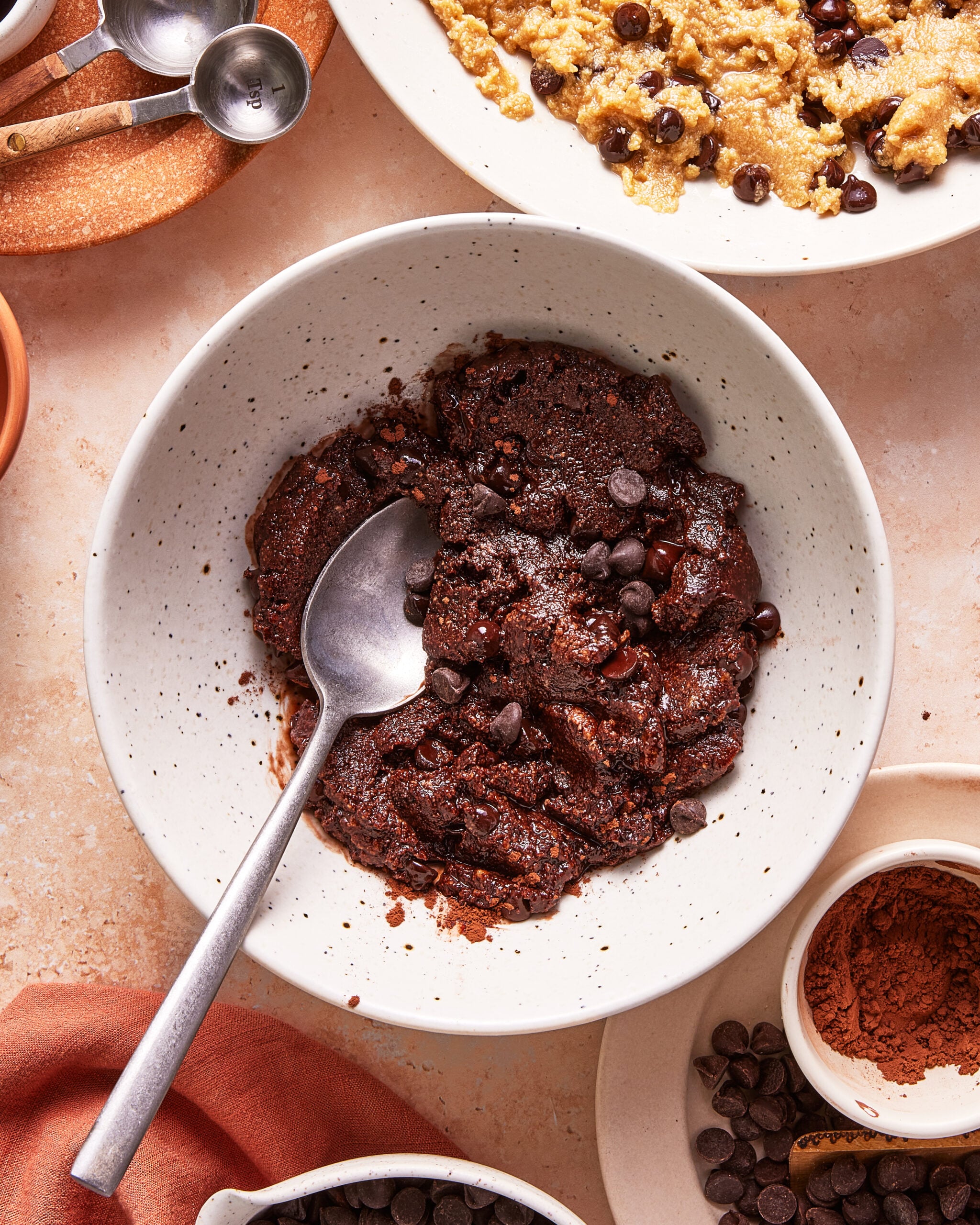 A bowl of chocolate cookie dough with chocolate chips and a spoon resting inside, surrounded by measuring spoons, scattered chocolate chips, cocoa powder, and another bowl of dough on a beige countertop.
