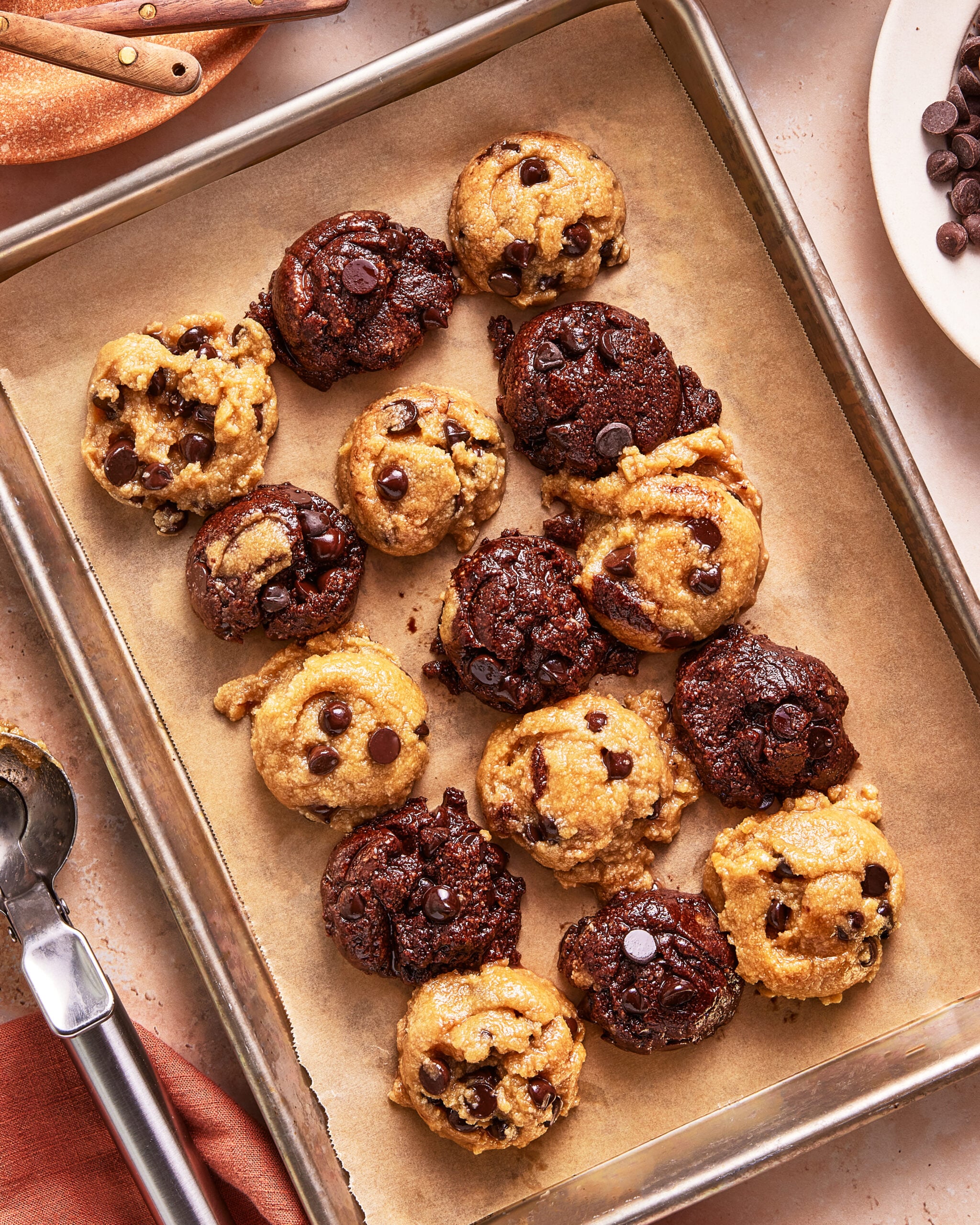 A baking sheet lined with parchment paper holds rows of freshly baked chocolate chip cookies—some golden brown, others dark chocolate—arranged in a mix of flavors. A scoop and spoon are nearby.