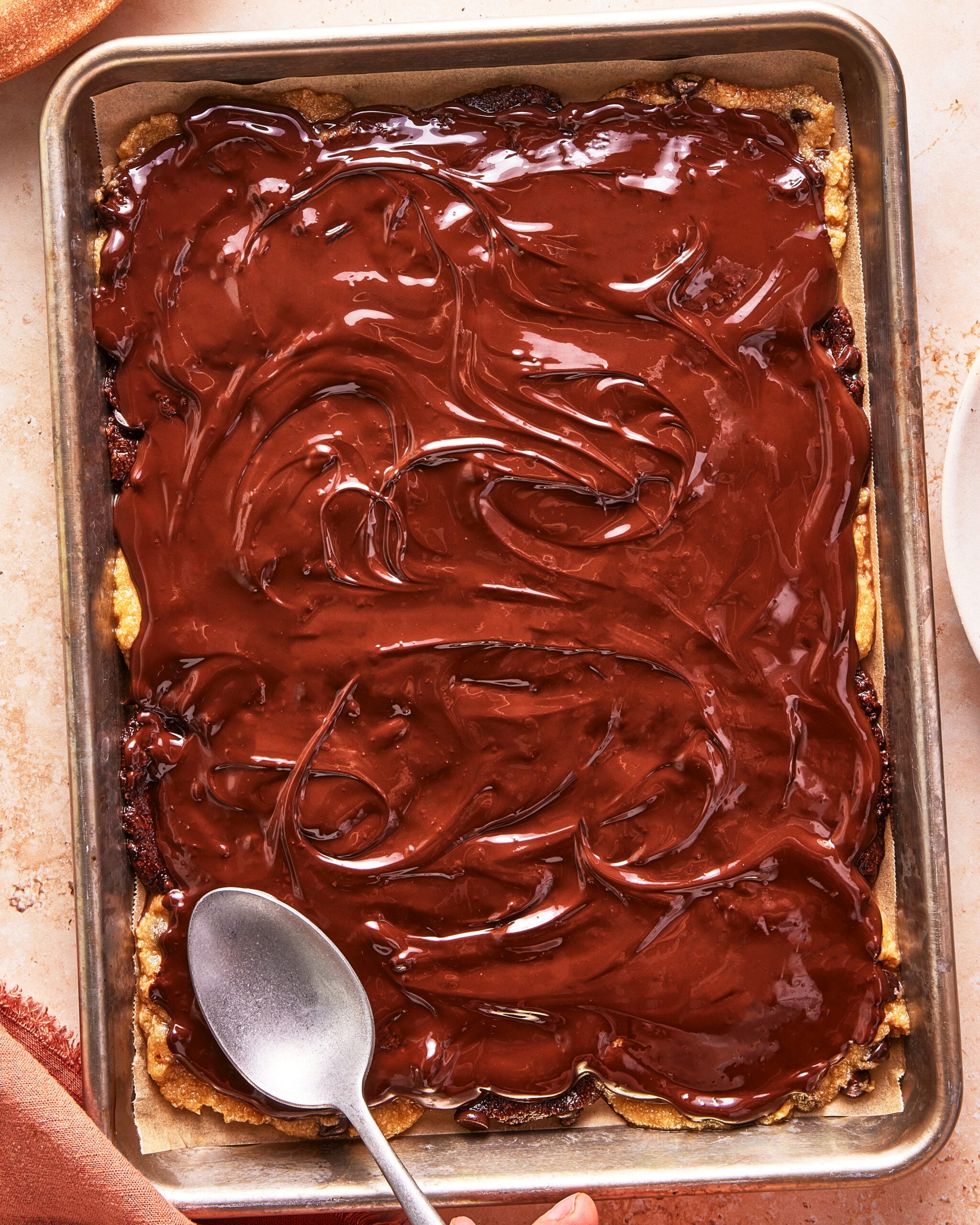 A rectangular baking tray filled with a dessert topped with a thick, glossy layer of melted chocolate, swirled with a spoon resting on one corner.