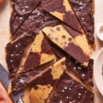 Chocolate-chip cookie and brownie bark broken into triangular pieces, covered with a layer of melted chocolate and sprinkled with flaky sea salt. A knife and part of a plate are visible beside the bark.