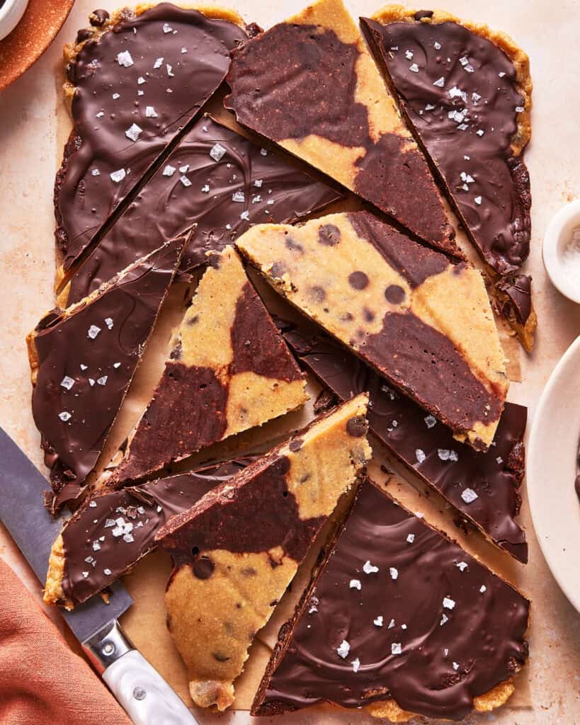 Chocolate-chip cookie and brownie bark broken into triangular pieces, covered with a layer of melted chocolate and sprinkled with flaky sea salt. A knife and part of a plate are visible beside the bark.