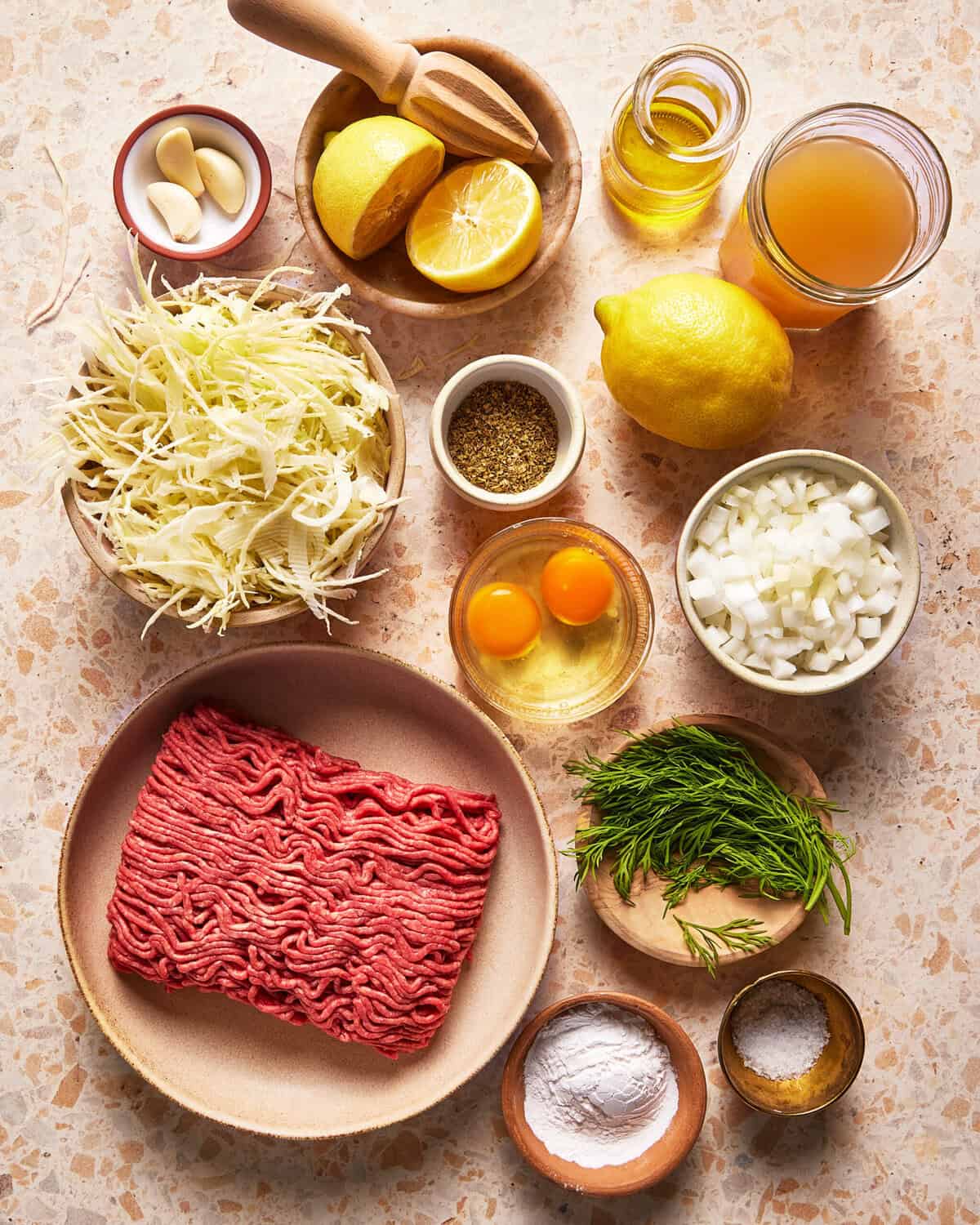 A top-down view of ingredients for a recipe, including ground meat, shredded cabbage, garlic, lemon, eggs, chopped onion, fresh dill, spices, flour, salt, olive oil, and broth, arranged on a light countertop.
