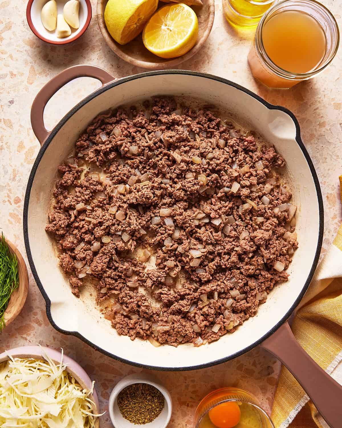 A skillet filled with browned ground beef and diced onions sits on a countertop, surrounded by ingredients like eggs, broth, sauerkraut, garlic, lemon, fresh herbs, and seasonings.