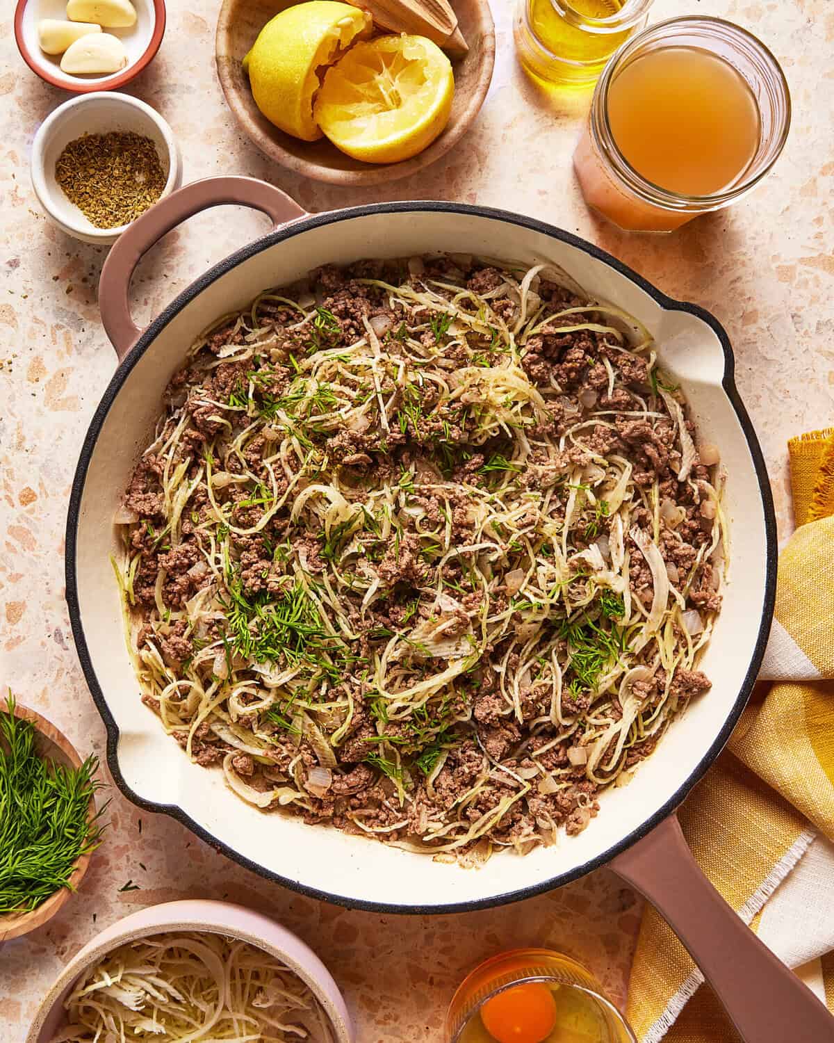A skillet filled with cooked ground meat, sliced onions, and fresh herbs sits on a countertop surrounded by bowls of spices, fresh dill, eggs, garlic, lemon, and a jar of broth.