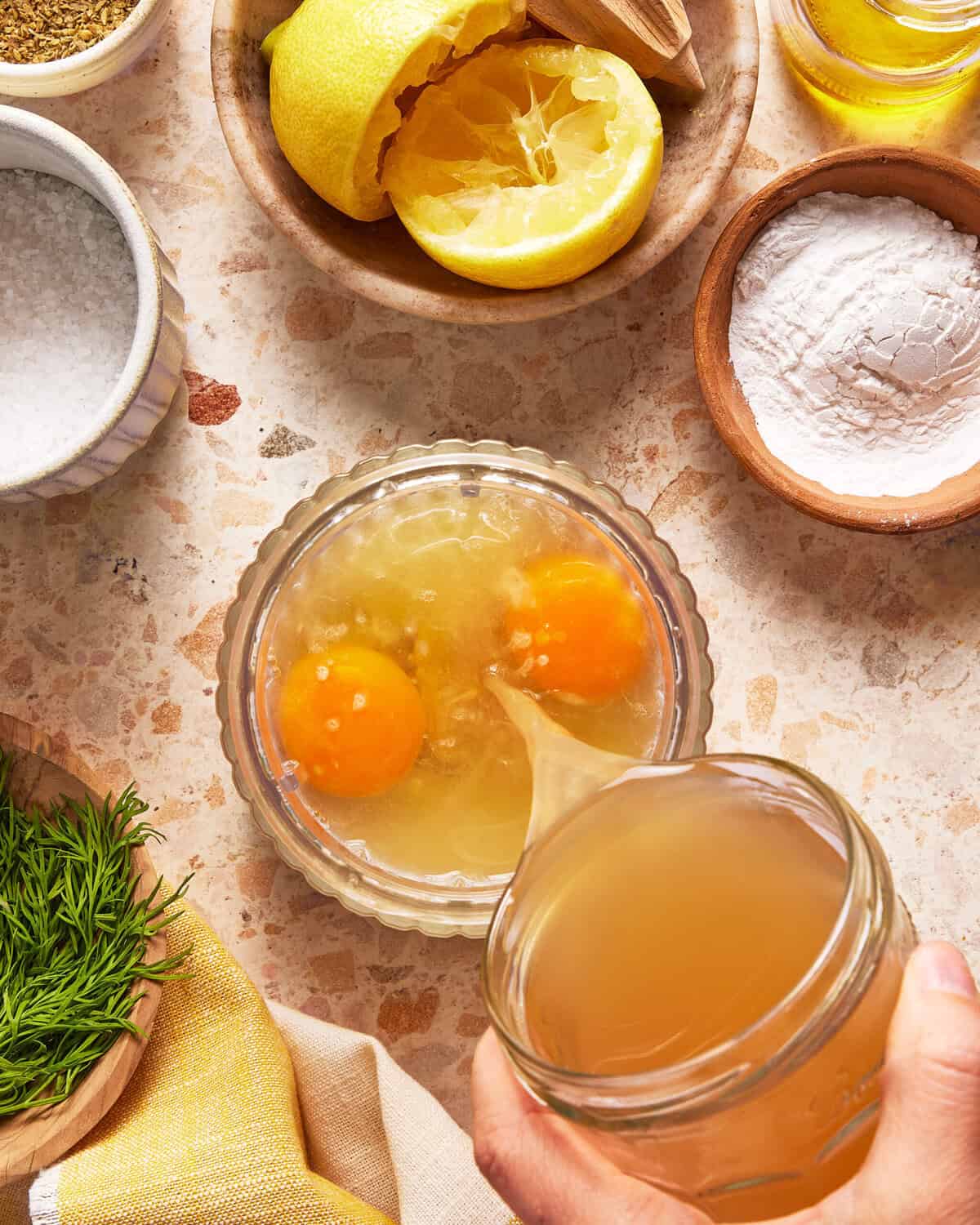 A hand pours broth into a bowl with two cracked eggs. Surrounding the bowl are ingredients: salt, flour, lemon halves, olive oil, and fresh dill on a beige speckled countertop.
