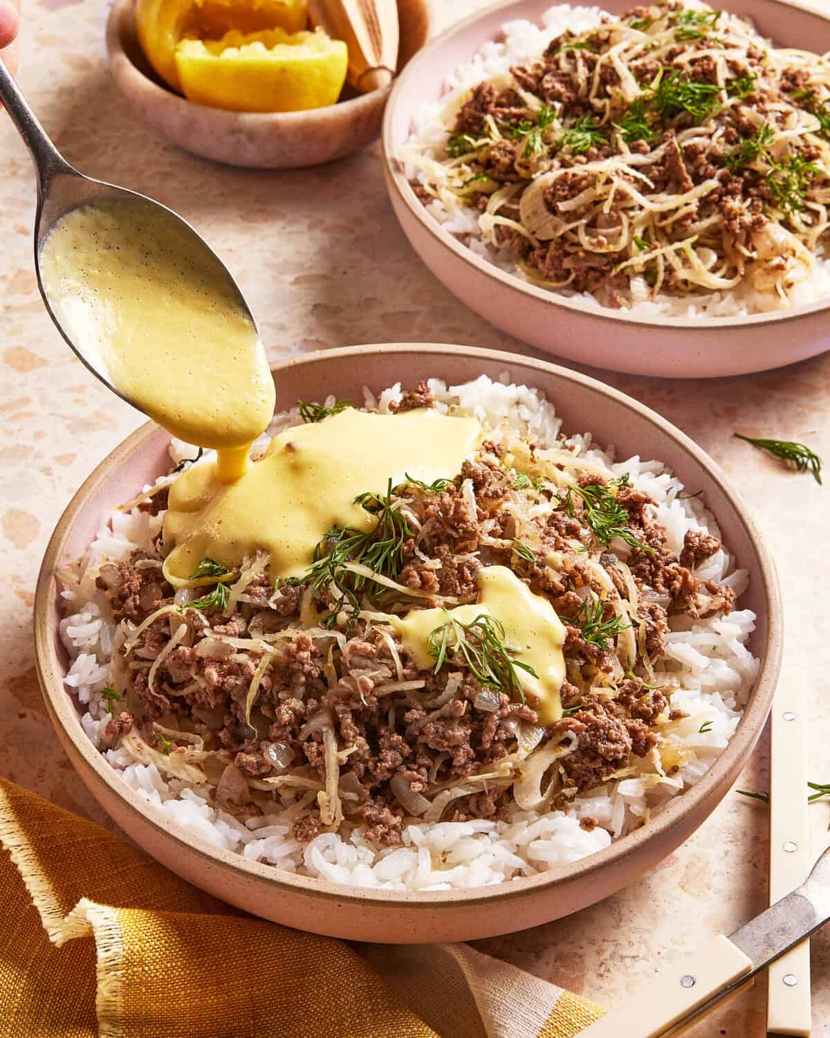 A spoon pours creamy yellow sauce over a bowl of rice topped with seasoned ground beef, herbs, and shredded cabbage. Another similar bowl and lemon wedges are visible in the background.