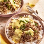 Two bowls filled with white rice, ground beef, caramelized onions, yellow sauce, and fresh dill. A glass of drink, a fork, and a yellow napkin are beside the bowls on a beige countertop.