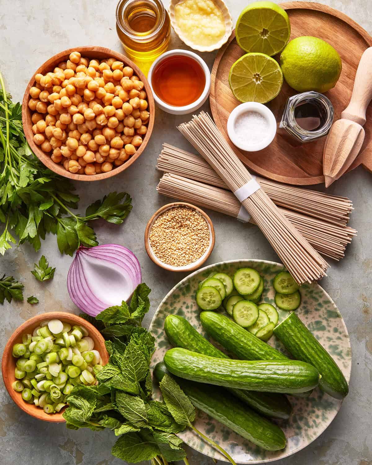 A flat lay of fresh ingredients including chickpeas, cucumbers, sliced red onion, green onions, mint, parsley, soba noodles, limes, garlic, sesame seeds, honey, soy sauce, and oil on a gray surface.