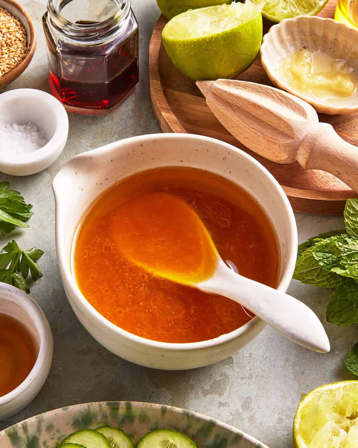 A bowl of orange sauce with a spoon inside, surrounded by lime halves, a wooden citrus reamer, mint leaves, a jar of liquid, and small bowls of salt and sesame seeds.