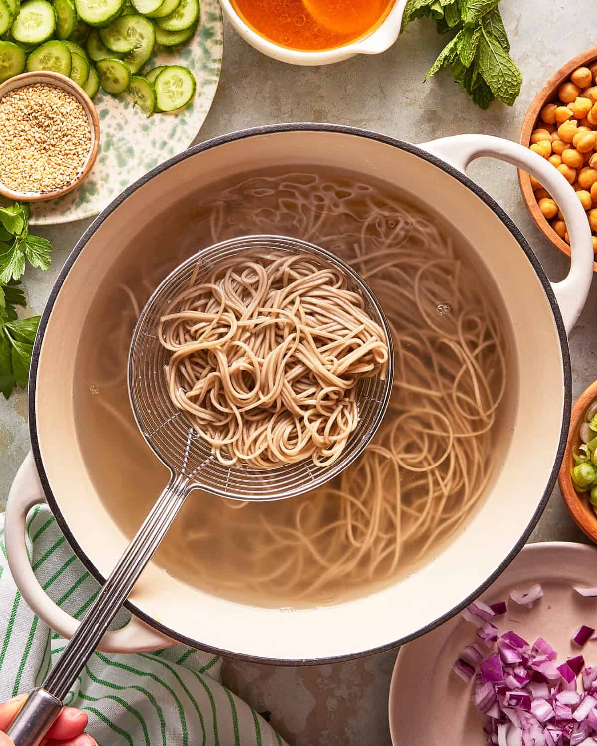 A large pot of cooked noodles in water sits on a table, surrounded by bowls and plates of fresh ingredients like sliced cucumbers, sesame seeds, chickpeas, chopped red onion, and leafy greens. A slotted ladle lifts noodles from the pot.