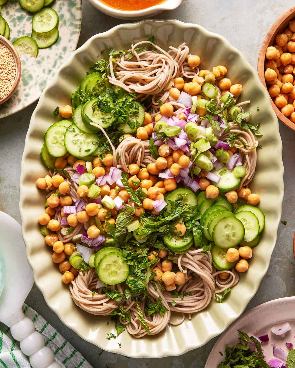 An oval plate filled with soba noodles, sliced cucumbers, chickpeas, chopped red onion, green onions, and fresh herbs, arranged in a colorful, fresh salad. Surrounding dishes contain dressing and sesame seeds.