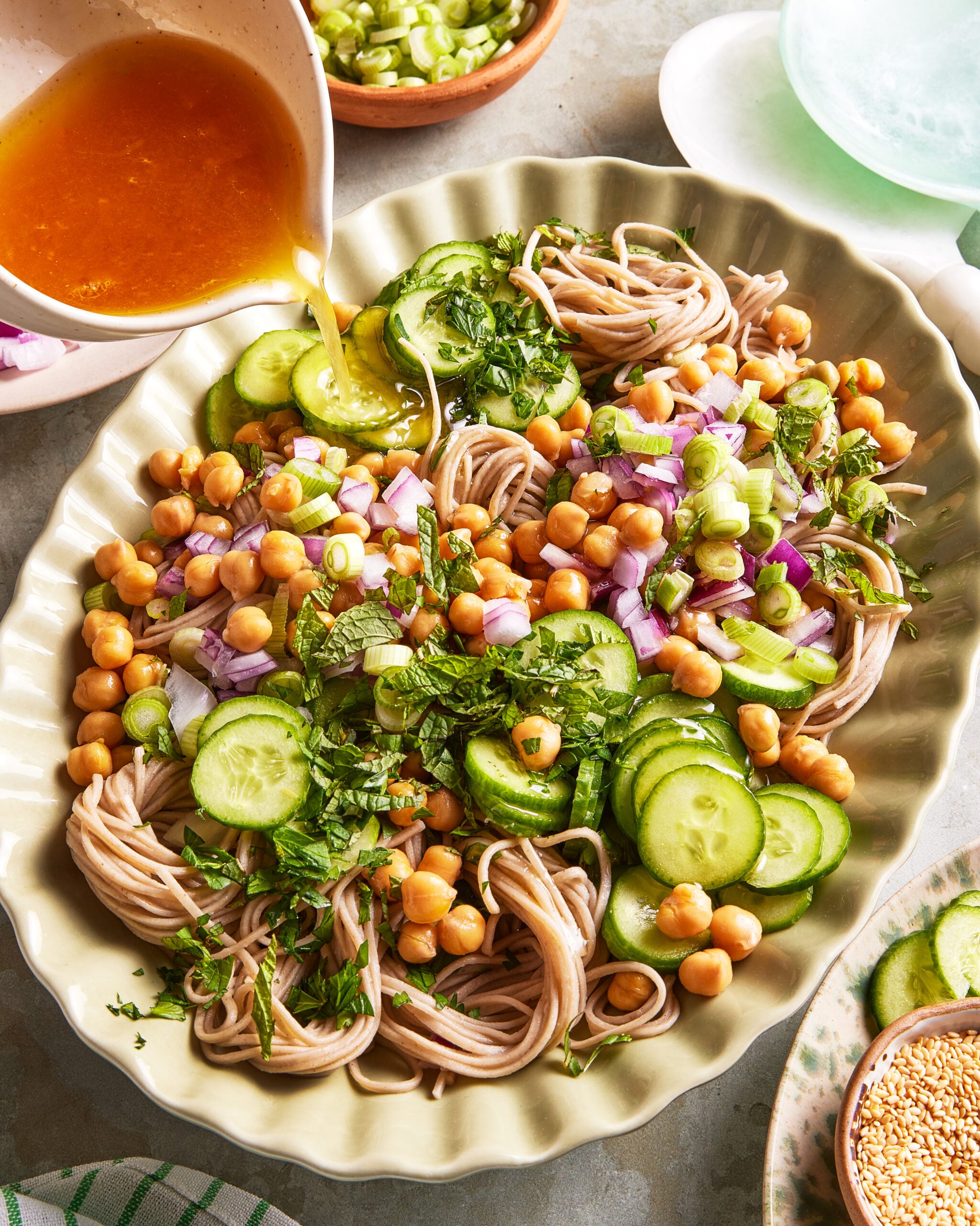 A platter of soba noodles topped with sliced cucumbers, chickpeas, chopped red onions, scallions, and herbs, with dressing being poured over the salad. Bowls of green onions and sesame seeds are nearby.