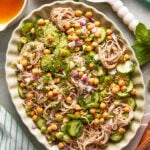 A large oval platter filled with soba noodles, sliced cucumbers, chickpeas, chopped red onion, fresh herbs, and sesame seeds, with a bowl of dressing and a bowl of sesame seeds nearby.