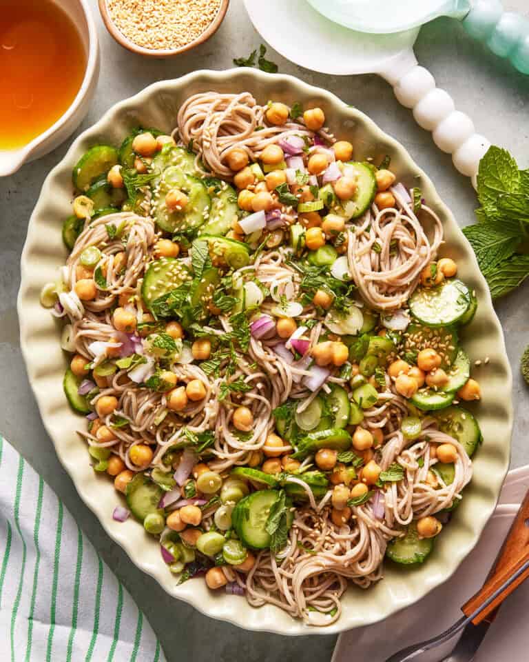 A large oval platter filled with soba noodles, sliced cucumbers, chickpeas, chopped red onion, fresh herbs, and sesame seeds, with a bowl of dressing and a bowl of sesame seeds nearby.