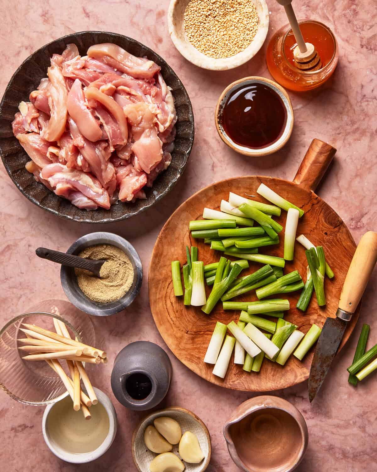 A top-down view of raw chicken strips, sliced green onions, garlic cloves, black pepper, soy sauce, sesame seeds, honey, and skewers arranged neatly on a pink marble surface with a knife.