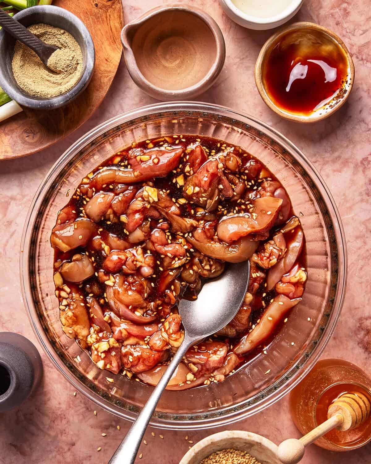 A glass bowl filled with raw chicken strips marinating in a dark sauce with garlic and sesame seeds, surrounded by small bowls of spices, sauce, honey, and sesame seeds on a pinkish countertop.