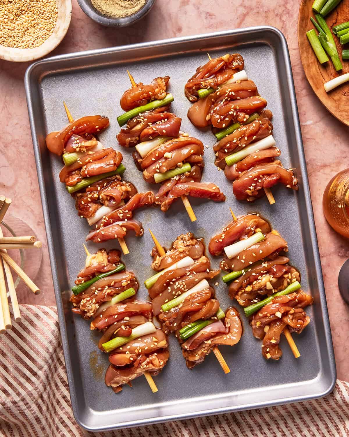 Raw chicken and green onion skewers arranged on a metal baking tray, sprinkled with sesame seeds and surrounded by small bowls of ingredients, wooden skewers, and a striped cloth on a pink surface.