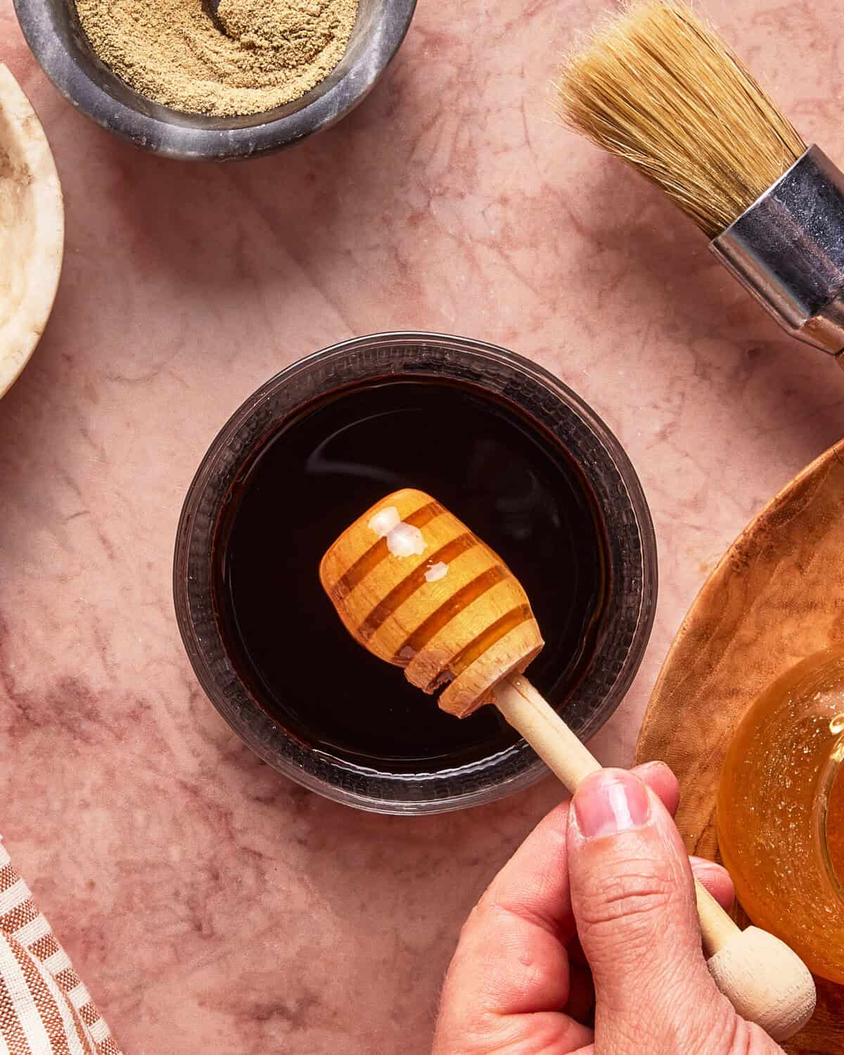 A hand holds a honey dipper over a bowl of dark liquid on a pink marble surface, surrounded by a brush, a bowl of powder, and a jar of honey.