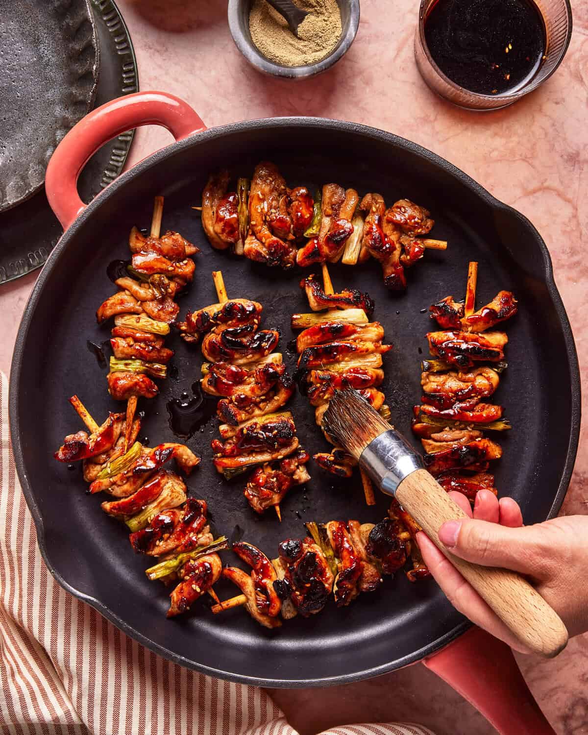A hand brushes glaze onto grilled chicken skewers with vegetables in a round black skillet, surrounded by bowls of seasoning and sauce on a pinkish countertop.