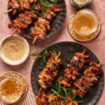 Two plates of grilled chicken skewers garnished with sesame seeds and green onions, surrounded by small bowls of sesame seeds, pepper, and drinks on a pink surface.