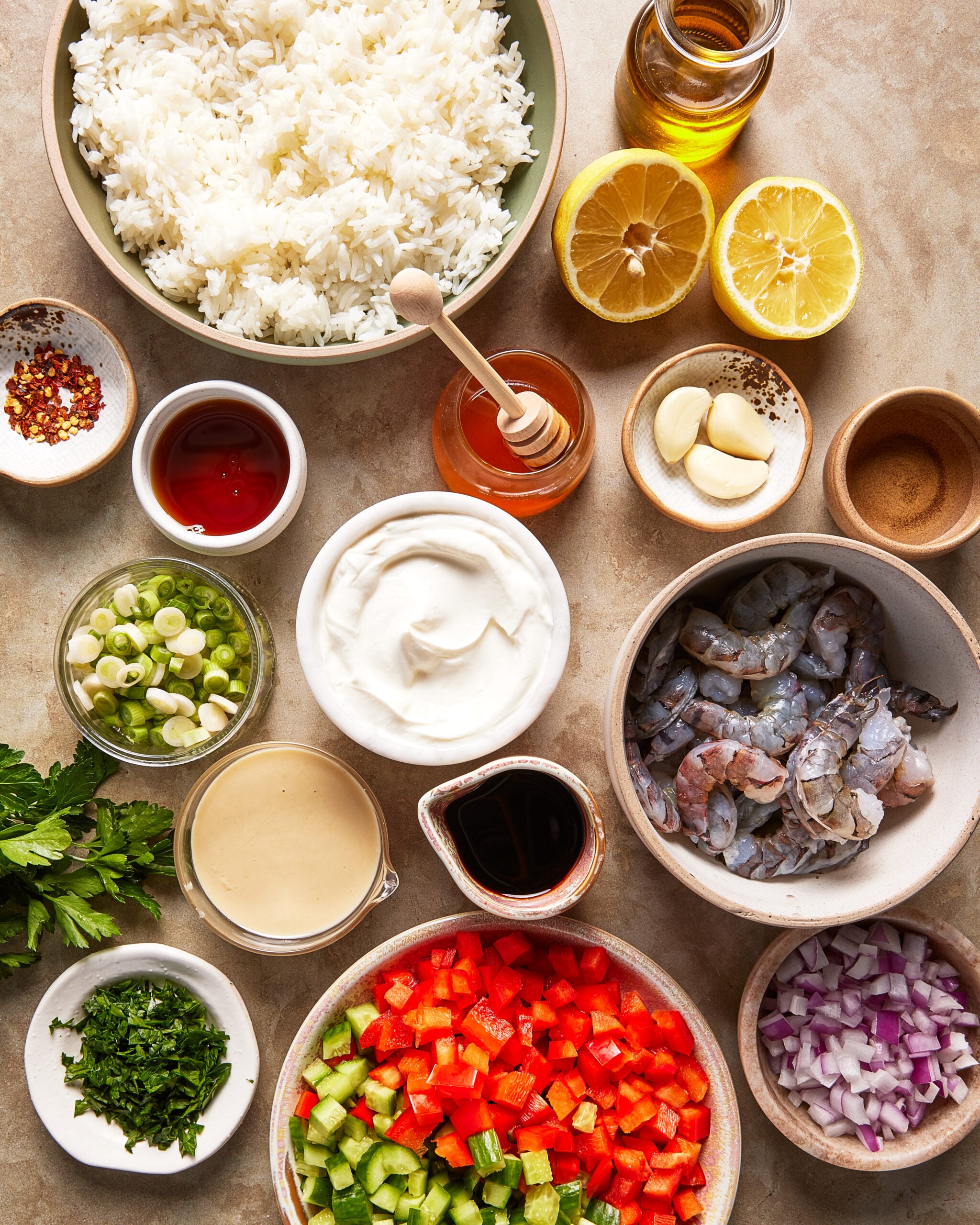 A top-down view of bowls containing cooked rice, raw shrimp, chopped red pepper, cucumber, red onion, green onion, fresh herbs, lemon halves, garlic cloves, yogurt, honey, tahini, soy sauce, chili flakes, oil, and vinegar.