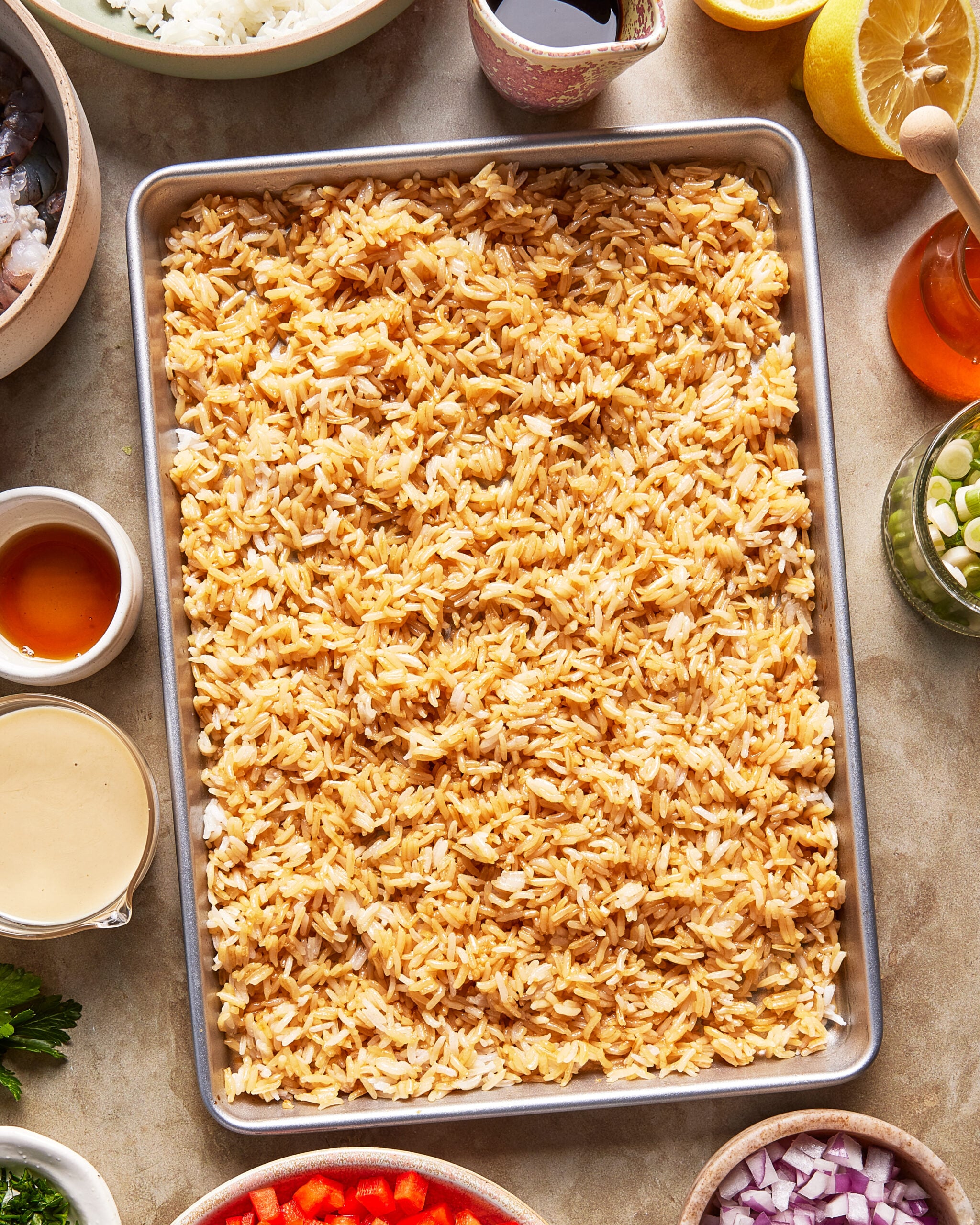 A sheet pan filled with browned rice, surrounded by small bowls of various ingredients including sauces, vegetables, herbs, lemon, and honey on a light surface.