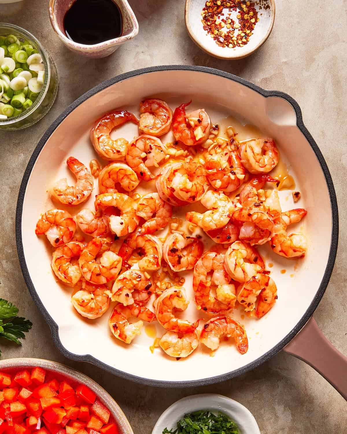 Shrimp sautéing in a white skillet with chopped garlic. Surrounding the skillet are bowls of sliced green onions, red pepper flakes, soy sauce, chopped red bell peppers, and fresh herbs.
