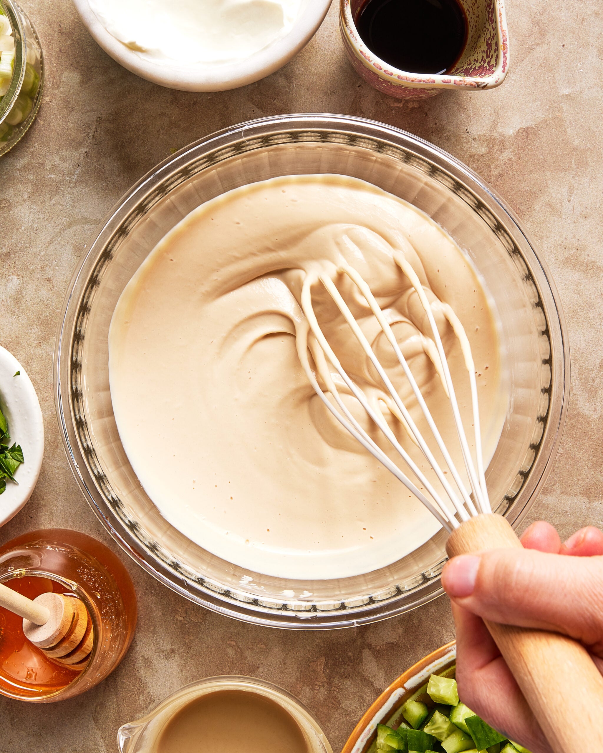 A hand holding a whisk is mixing a creamy light brown sauce in a glass bowl, surrounded by bowls of honey, chopped vegetables, yogurt, and a dark liquid on a countertop.