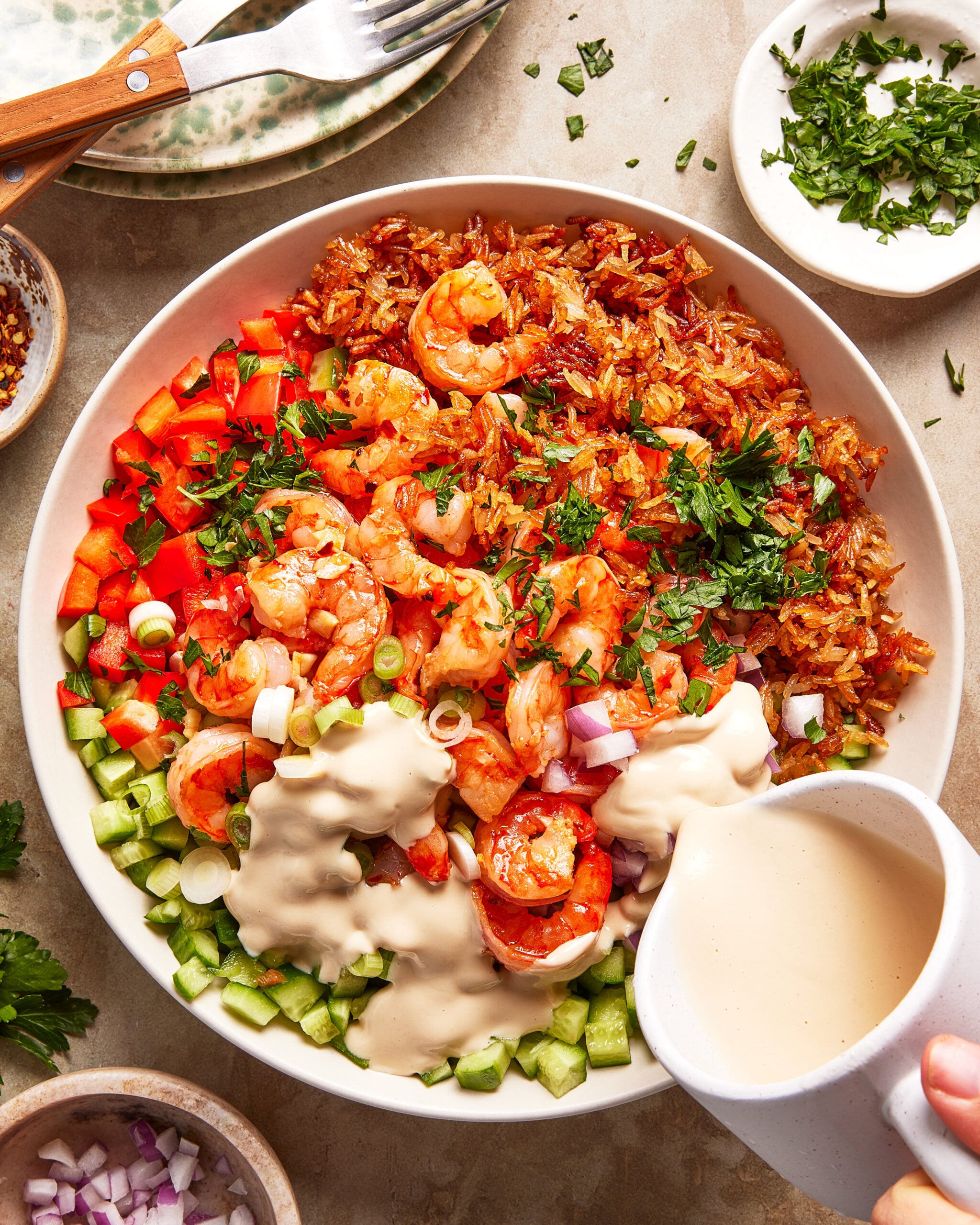 A bowl filled with shrimp, chopped red bell pepper, cucumber, rice, and fresh herbs, with a creamy sauce being poured over, surrounded by small bowls of chopped herbs and a plate with utensils.