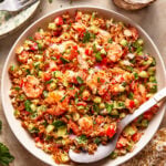 A bowl of colorful fried rice with shrimp, diced bell peppers, scallions, and fresh herbs. A large serving spoon rests in the bowl, and small plates and glasses are visible nearby on a beige tabletop.