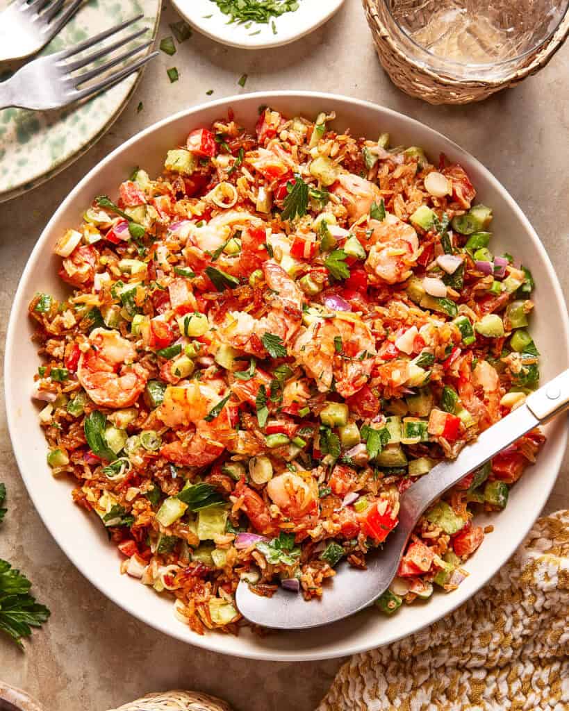 A bowl of colorful fried rice with shrimp, diced bell peppers, scallions, and fresh herbs. A large serving spoon rests in the bowl, and small plates and glasses are visible nearby on a beige tabletop.