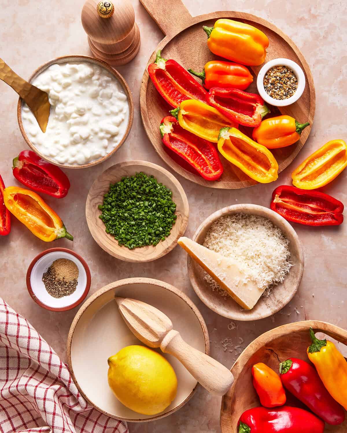 A top view of colorful mini bell peppers, cottage cheese, grated Parmesan, chopped chives, black pepper, a lemon, a citrus reamer, a pepper mill, and a red-striped towel arranged on a beige surface.