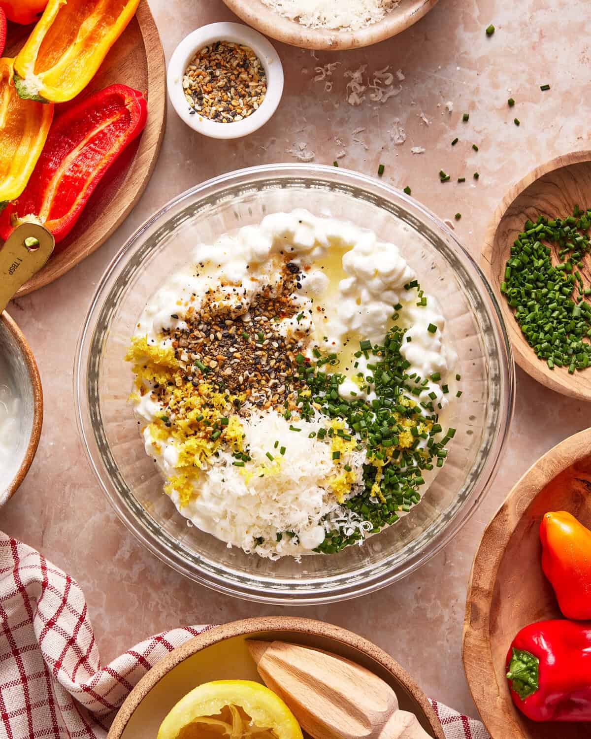 A glass bowl with cottage cheese, grated cheese, chopped chives, lemon zest, pepper, and seasonings sits on a counter surrounded by peppers, chives, lemon, and small wooden bowls of ingredients.