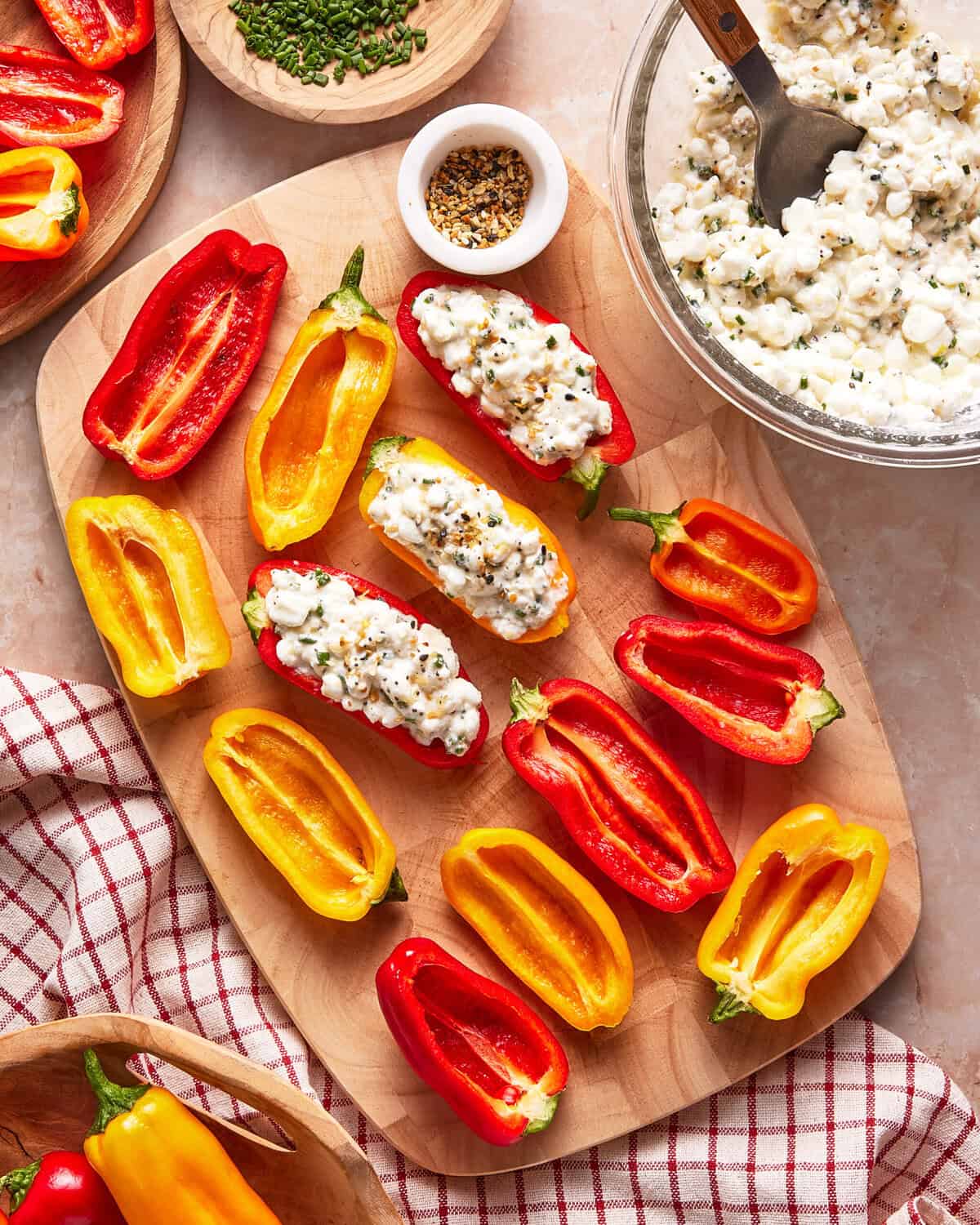 A wooden board with halved red and yellow mini bell peppers, some stuffed with cottage cheese and seasonings. A small bowl of spices, a bowl of cottage cheese, and a red-checked cloth are nearby.