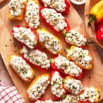 Mini bell peppers halved and filled with a creamy cottage cheese mixture, garnished with chopped chives and seasoning, arranged on a wooden cutting board with a small bowl of spices nearby.