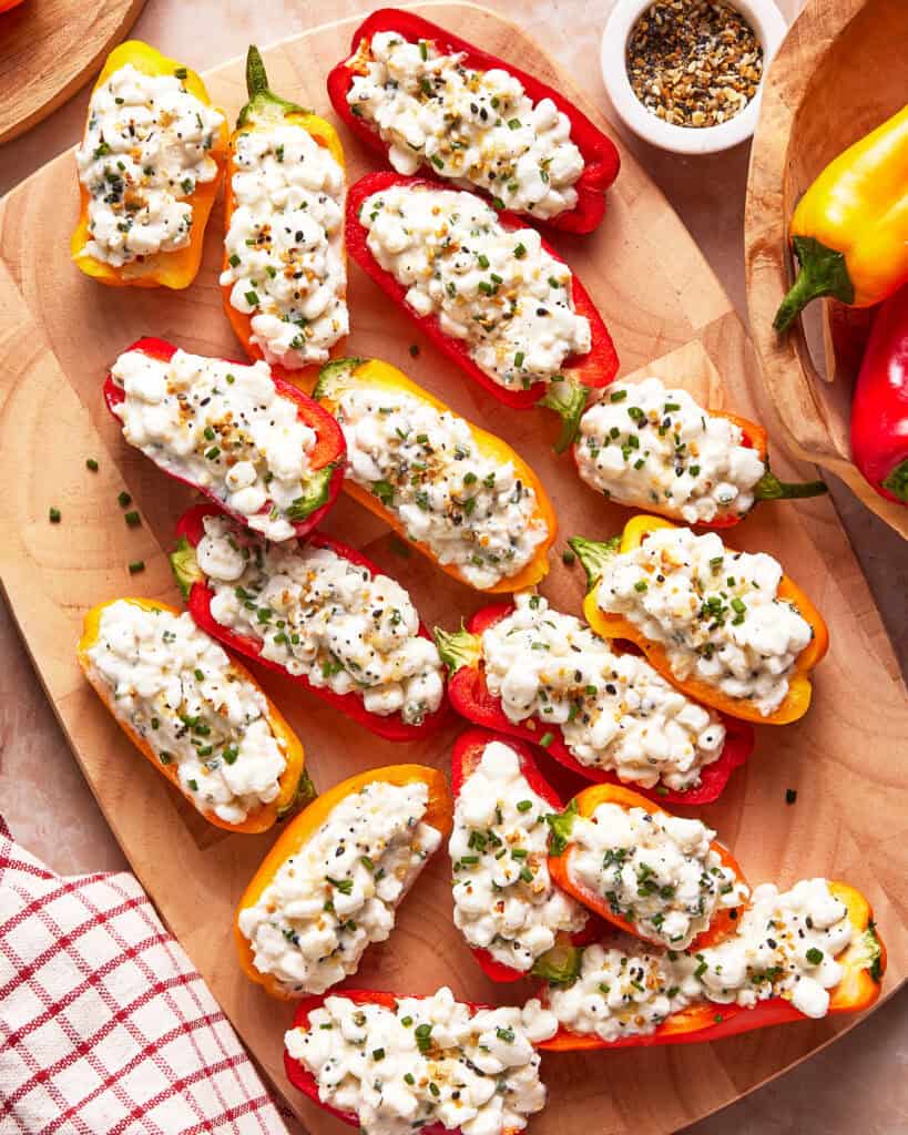 Mini bell peppers halved and filled with a creamy cottage cheese mixture, garnished with chopped chives and seasoning, arranged on a wooden cutting board with a small bowl of spices nearby.