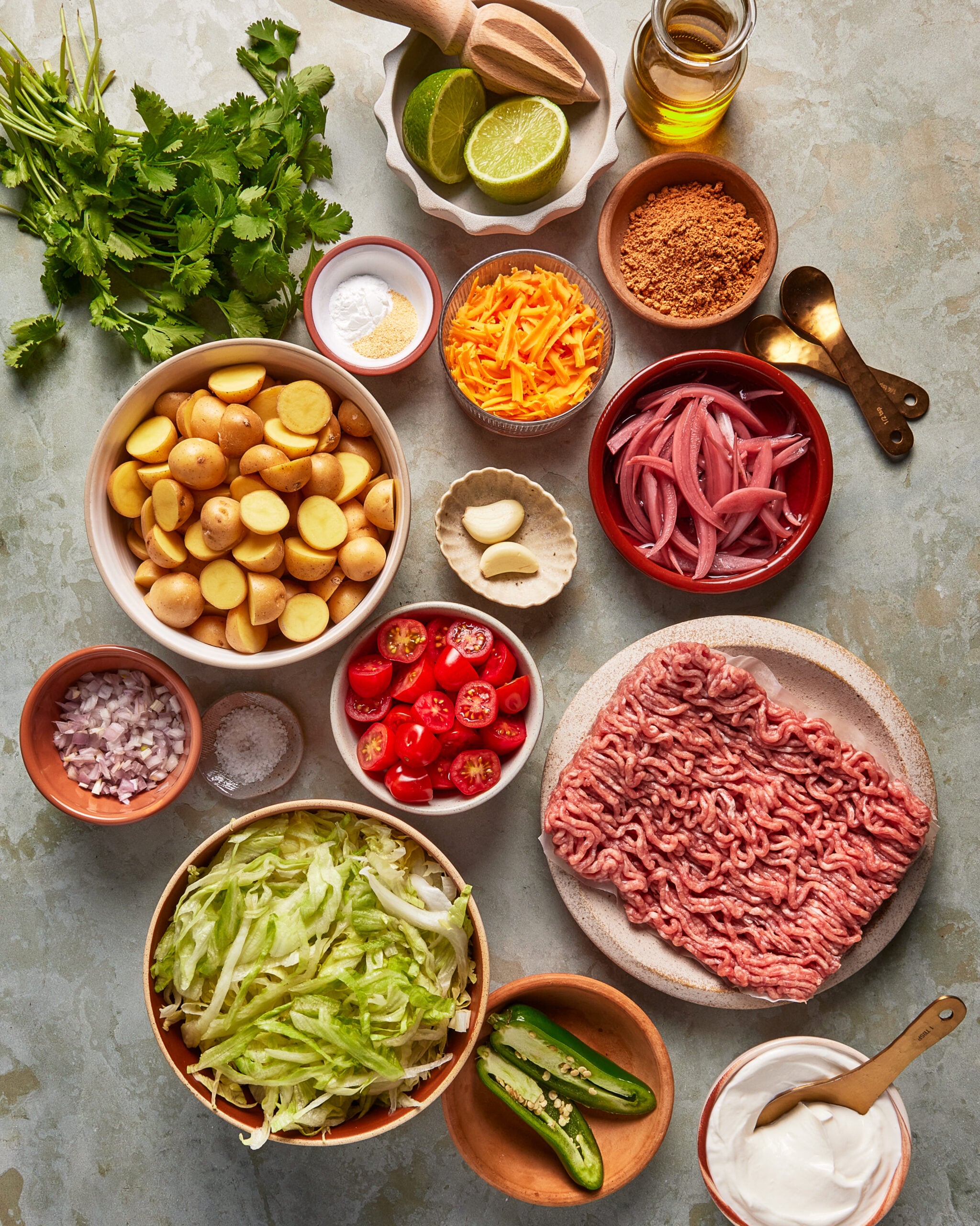 A variety of taco ingredients arranged on a counter, including ground meat, lettuce, baby potatoes, shredded cheese, cherry tomatoes, pickled onions, jalapeños, garlic, limes, seasonings, cilantro, and a jar of oil.
