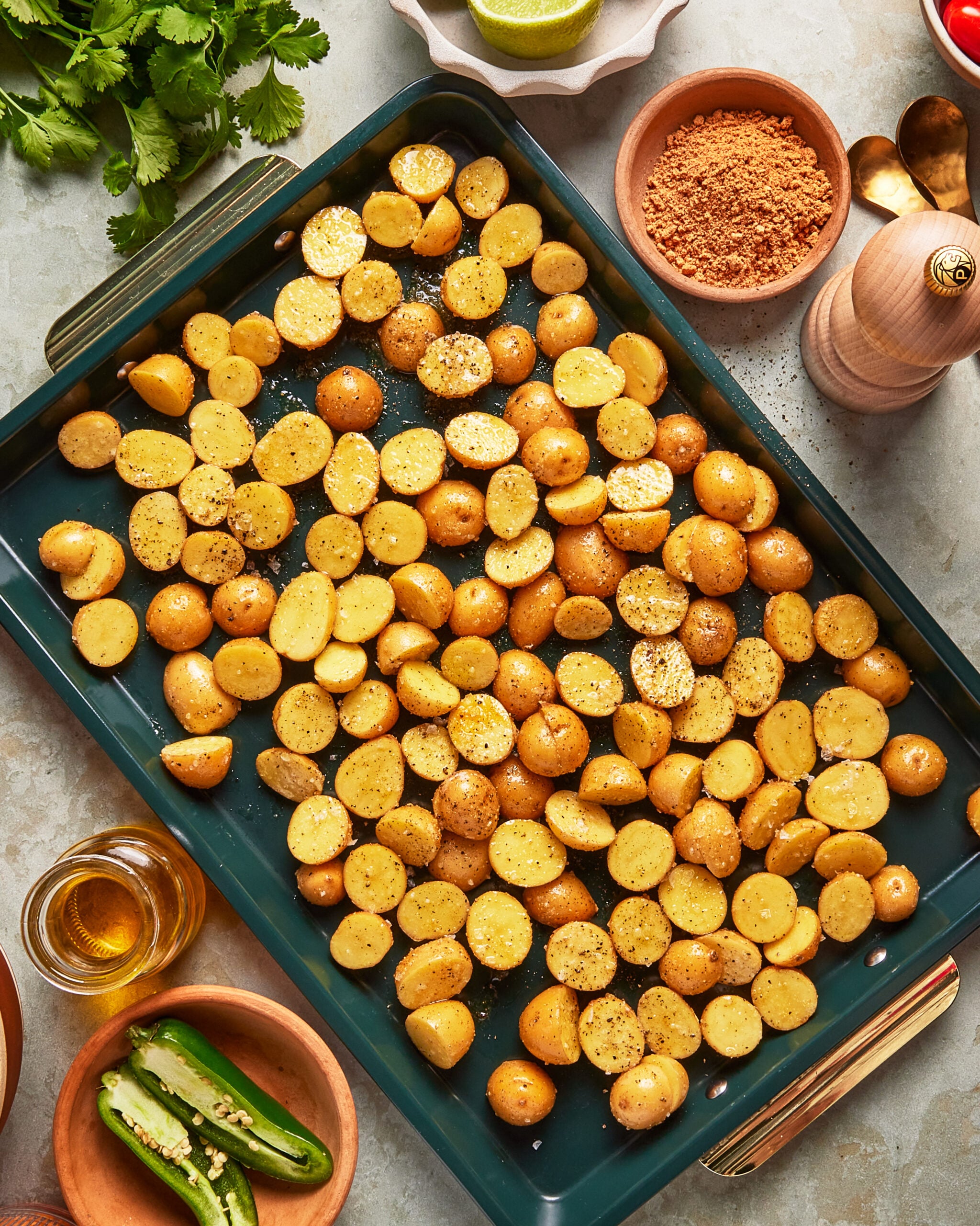 A baking tray filled with halved baby potatoes seasoned with pepper and salt, surrounded by ingredients including sliced jalapeños, cilantro, lime, a bowl of spices, olive oil, tomatoes, and a pepper grinder.