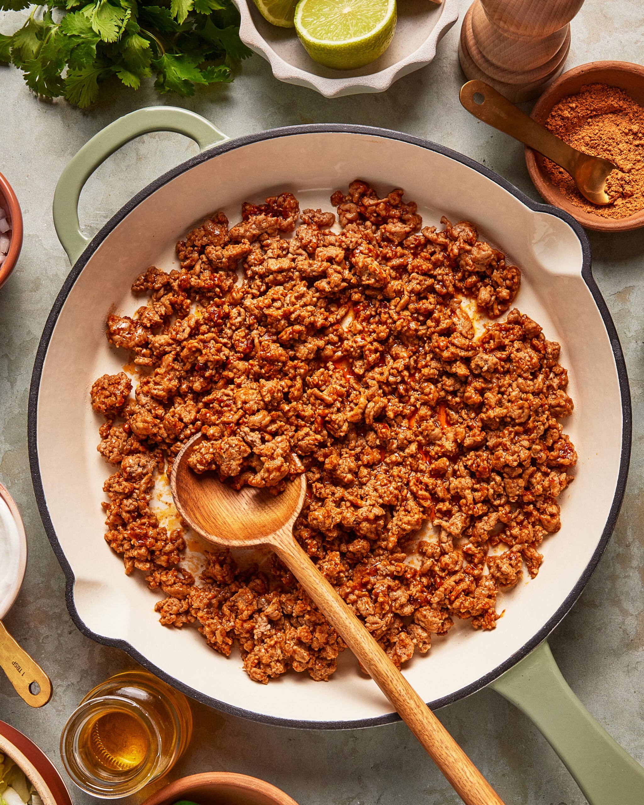 Ground meat cooked with seasoning in a large skillet, stirred with a wooden spoon. Surrounding the skillet are bowls of spices, chopped onions, oil, fresh cilantro, and halved limes on a countertop.