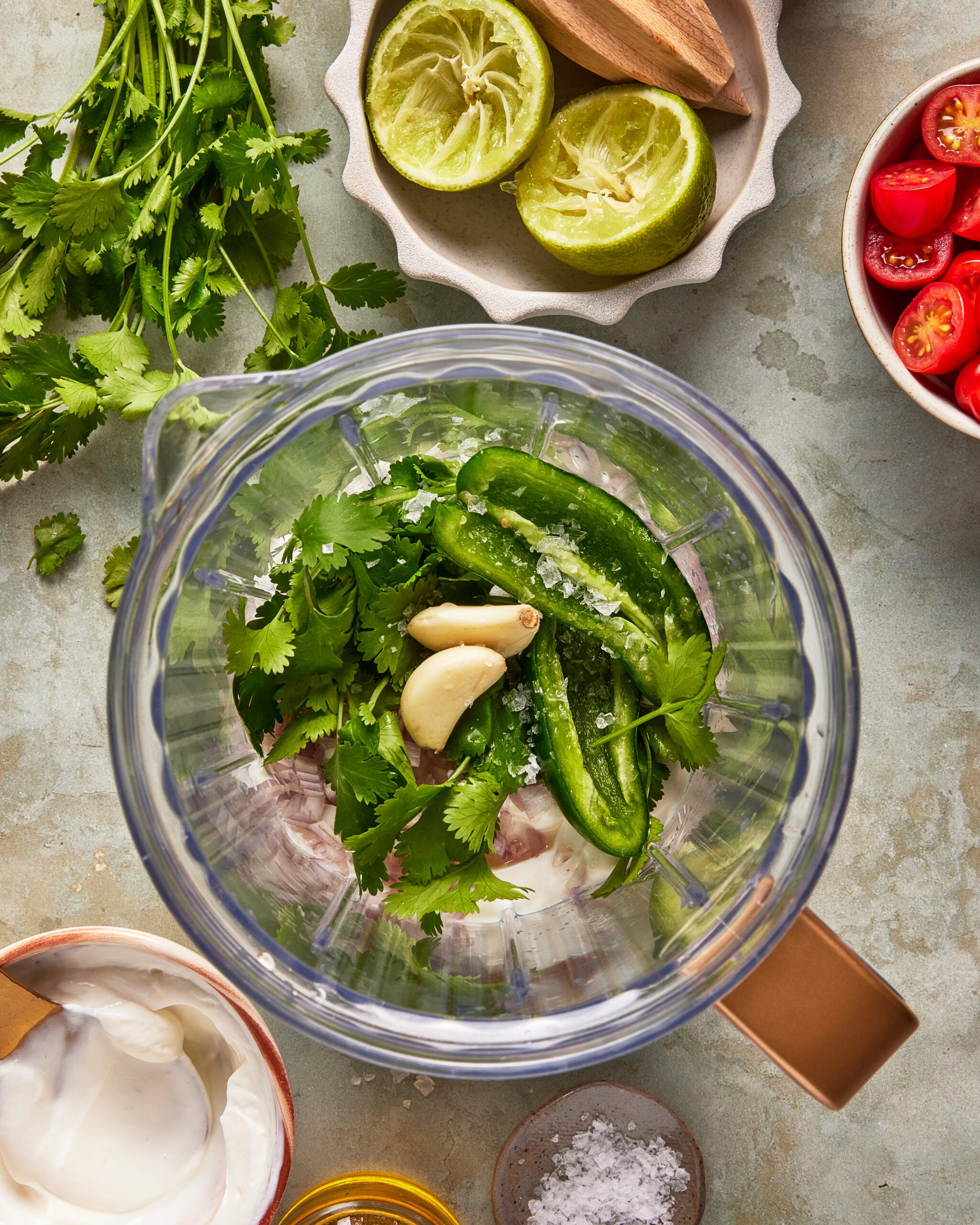 Overhead view of a blender with cilantro, jalapeño, garlic, and salt inside. Surrounding the blender are halved limes, cherry tomatoes, fresh cilantro, a bowl of sour cream, a small bowl of salt, and a small bowl of oil.