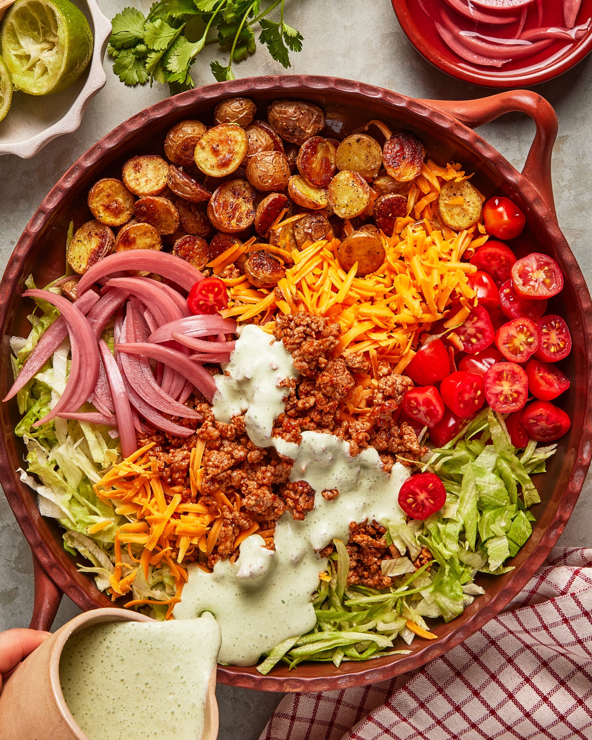 A large bowl filled with shredded lettuce, halved cherry tomatoes, grated cheddar cheese, sliced pickled onions, crispy roasted potatoes, seasoned ground meat, and creamy dressing being poured over the top.